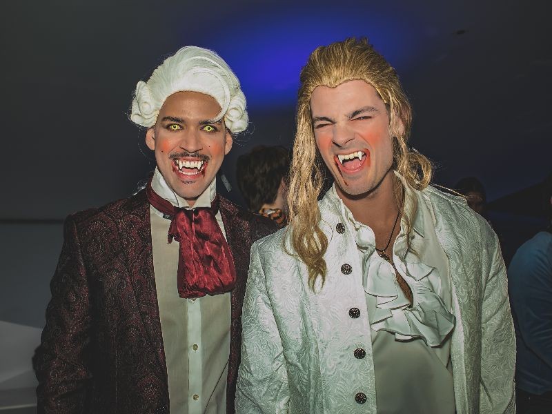 Two people in vampire costumes with powdered wigs, white face makeup and fangs, smiling at a party