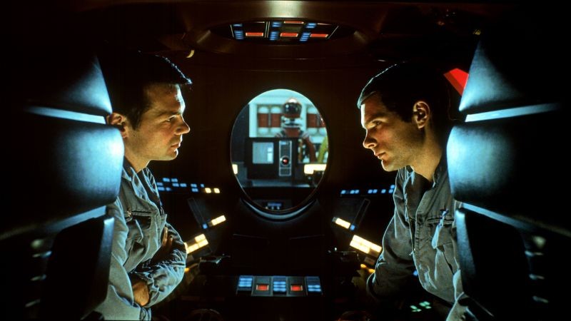 Two astronauts in a dim spacecraft pod lit by red and blue control panels