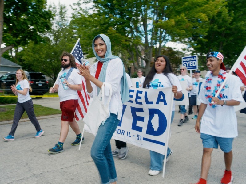 People marching down a suburban street carrying American flags and blue campaign signs