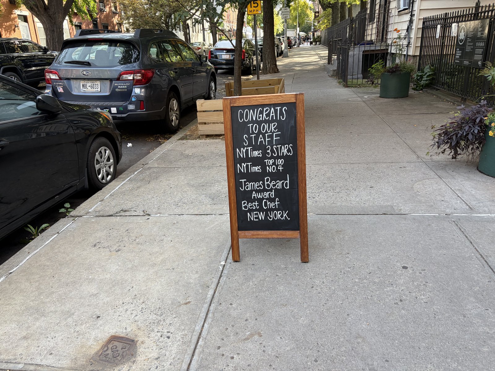 A chalkboard on the sidewalk reads "Congrats to our staff / NYTimes 3 stars / NYTimes Top 100 No. 4 / James Beard Award Best Chef New York." Parked cars and street trees line the block.