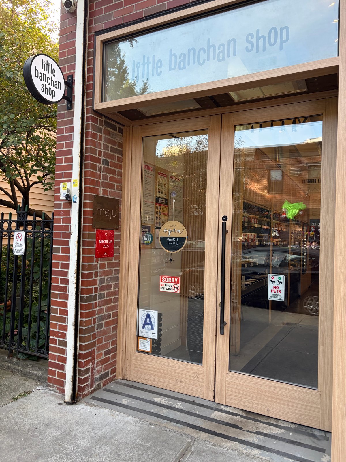 Wood-framed glass doors under a "little banchan shop" sign. A "meju" plaque and a red "Michelin 2025" plaque hang on the brick wall. Signs on the door show the hours (open 10 to 9, Sunday 10 to 8), an NYC Health "A" grade, a Beli sticker, "No public restrooms" and "No pets."