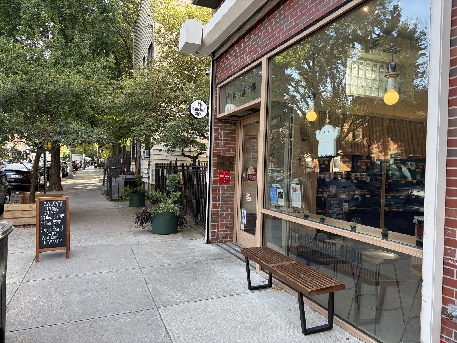 A brick storefront with large windows and a "little banchan shop" sign on a tree-lined sidewalk. A wooden bench is in front, and a sidewalk chalkboard stands to the left.